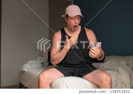 Surprised Caucasian Man in Pink Cap Looking at Smartphone on Bed in Bedroom Surprised Caucasian Man in Pink Cap Looking at Smartphone on Bed in Bedroom 138451852