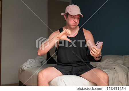 Caucasian Man in Pink Cap Giving Thumbs Down to Smartphone on Bed in Bedroom 138451854