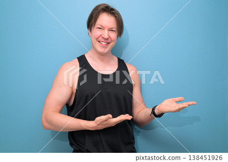 Smiling Caucasian Man in Black Tank Top Gesturing Sideways on Blue Background Smiling Caucasian Man in Black Tank Top Gesturing Sideways on Blue Background 138451926