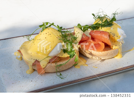 Two perfectly poached eggs rest atop toasted English muffins, draped in a rich, velvet-smooth hollandaise sauce that glistens under soft light. Nestled beneath the eggs are delicate ribbons of smoked 138452331