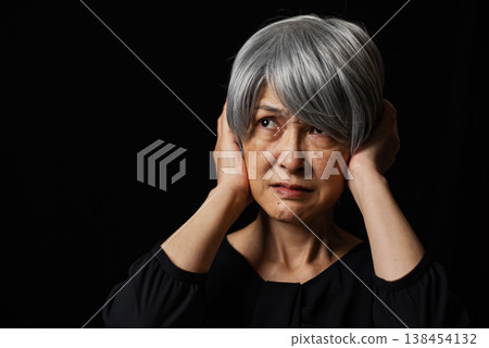 An image of a senior woman holding her head in her hands in front of a black background. 138454132
