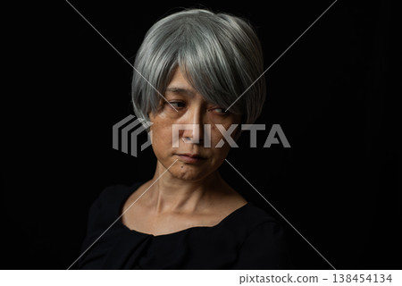 A senior woman with white hair glares resentfully in front of a black background. 138454134