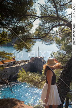 Woman in summer skirt and straw hat spins with open arms on cliff above rocks Adriatic sea and green hills Montenegro travel inspiration scene Woman in summer skirt and straw hat spins with open arms on cliff above rocks Adriatic sea and green hills Montenegro travel inspiration scene 138454270