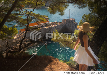 Woman in summer skirt and straw hat spins with open arms on cliff above rocks with Adriatic waves and green hills Montenegro travel scene Woman in summer skirt and straw hat spins with open arms on cliff above rocks with Adriatic waves and green hills Montenegro travel scene 138454271