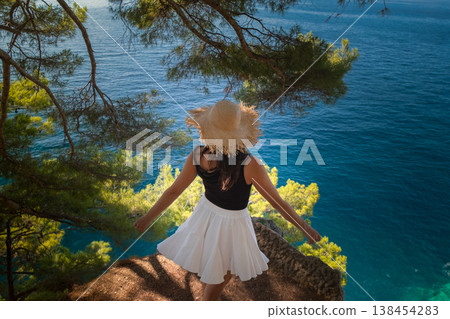 Woman in summer skirt and straw hat spins and leans on cliff above rocks Adriatic waves and green hills Montenegro travel freedom vibe Woman in summer skirt and straw hat spins and leans on cliff above rocks Adriatic waves and green hills Montenegro travel freedom vibe 138454283