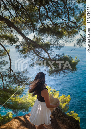 Back view of girl in summer outfit with flowing skirt and hat twirling skirt on cliff with Adriatic waves crashing and green Montenegro hills background Back view of girl in summer outfit with flowing skirt and hat twirling skirt on cliff with Adriatic waves crashing and green Montenegro hills background 138454284