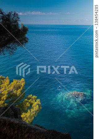 View of rocky Adriatic coast framed by green trees with blue water hitting stones and green mountains in background View of rocky Adriatic coast framed by green trees with blue water hitting stones and green mountains in background 138454285