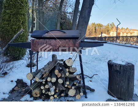 Winter, in the yard with a metal barbecue with a lid and stacked firewood under it. Snow, bare trees, an atmosphere of comfort and preparation for cooking outdoors. Countryside vacation, barbecue Winter, in the yard with a metal barbecue with a lid and stacked firewood under it. Snow, bare trees, an atmosphere of comfort and preparation for cooking outdoors. Countryside vacation, barbecue 138461298