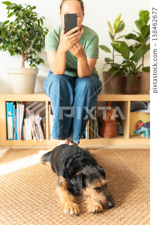 Woman sitting on a shelf, focused on smartphone, with her small cute pet sitting on a rug nearby at home. dog. An authentic moment of life Woman sitting on a shelf, focused on smartphone, with her small cute pet sitting on a rug nearby at home. dog. An authentic moment of life 138462677
