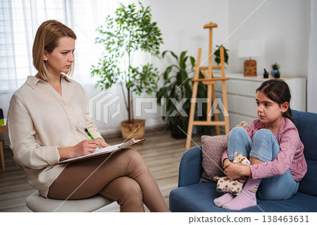 Psychologist writing notes while young girl sitting on sofa Psychologist writing notes while young girl sitting on sofa 138463631