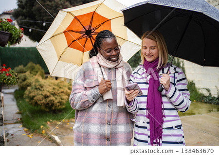 Diverse women friends smiling using smartphone under umbrellas in rain Diverse women friends smiling using smartphone under umbrellas in rain 138463670
