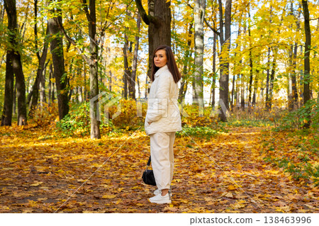 Successful woman walking in autumn forest. Caucasian businesswoman dressed in white quilted jacket enjoying nature. Blogger and mother relaxing outdoors during sunny fall season. 138463996