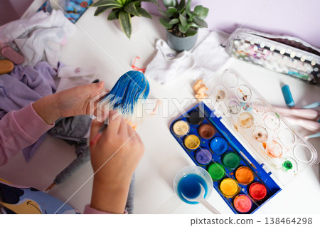 A childs hands paint a dolls hair blue with a brush on a table with a paint cup and palette surrounded by plants and art supplies creative hobby crafting diy concept A childs hands paint a dolls hair blue with a brush on a table with a paint cup and palette surrounded by plants and art supplies creative hobby crafting diy concept 138464298