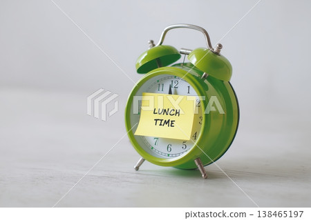 Green alarm clock displays a reminder for lunch time on a sticky note against a neutral backdrop. Green alarm clock displays a reminder for lunch time on a sticky note against a neutral backdrop. 138465197