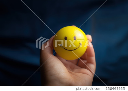 Hand holds yellow smiley face ball against a dark blue backdrop radiating a sense of optimism and cheerfulness. 138465635