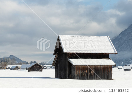 Snow covered wooden barn in Bavarian winter field with mountains and dramatic clouds Snow covered wooden barn in Bavarian winter field with mountains and dramatic clouds 138465822