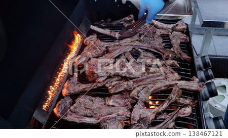 Chef wearing blue gloves uses tongs to grill lamb chops on a barbecue grill during an outdoor barbecue party, flames licking the meat as it cooks Chef wearing blue gloves uses tongs to grill lamb chops on a barbecue grill during an outdoor barbecue party, flames licking the meat as it cooks 138467121