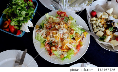 Caesar salad featuring grilled chicken, crisp croutons, shaved parmesan, and ripe tomatoes resting on rustic wooden surface near complementary restaurant dishes 138467126