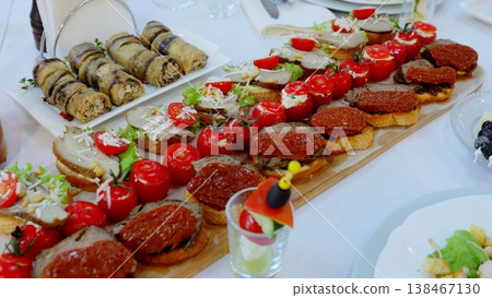 Rustic wooden tray displaying gourmet appetizers, including mini sandwiches, grilled eggplant rolls, cherry tomatoes, and charcuterie spread on crisp white linen at catered gathering Rustic wooden tray displaying gourmet appetizers, including mini sandwiches, grilled eggplant rolls, cherry tomatoes, and charcuterie spread on crisp white linen at catered gathering 138467130