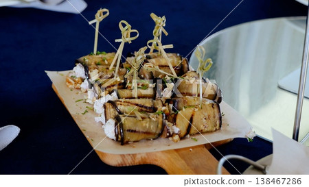 Grilled eggplant rolls stuffed with creamy cheese, toasted nuts, aromatic herbs, plated on rustic wooden board with garnishing skewers Grilled eggplant rolls stuffed with creamy cheese, toasted nuts, aromatic herbs, plated on rustic wooden board with garnishing skewers 138467286