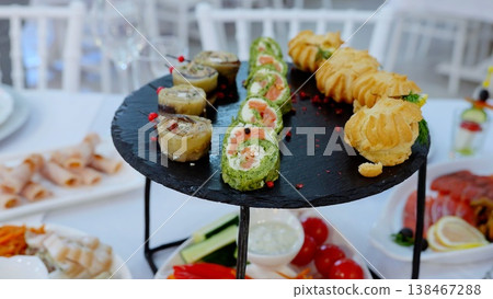 Slate round plate displaying elegant appetizers like pinwheel sandwiches, eclairs, and vegetable rolls, enhancing sophisticated catering presentation for festive gathering Slate round plate displaying elegant appetizers like pinwheel sandwiches, eclairs, and vegetable rolls, enhancing sophisticated catering presentation for festive gathering 138467288