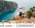 Young woman standing on a rock cliff and waving the US flag while looking at sea beneath. Girl traveller waving the American flag while standing on a mountain top. 4 fourth July Independence Day. 138468465