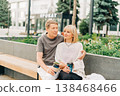 An adult mature happy couple in love sitting on bench outdoors in city street park. A blonde caucasian man and woman spend time together and drinking coffee. Senior wife and husband walking outside. 138468466