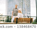 An adult mature happy romantic couple in love sitting on bench outdoors in city street park. A blonde caucasian man and woman spend time together. Senior wife and husband walking outside. 138468470