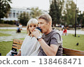 An adult mature happy couple in love sitting on bench outdoors in city street park. A blonde caucasian man and woman spend time together and drinking coffee. Senior wife and husband walking outside. 138468471