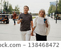 Adult mature happy romantic couple in love outdoors in city street park. A blonde caucasian man and woman spend time together, drinking coffee. Senior wife and husband walking outside on town square. 138468472