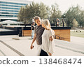 An adult mature happy romantic couple in love outdoors in city street park. A blonde caucasian man and woman spend time together. Senior wife and husband walking outside on town square. 138468474