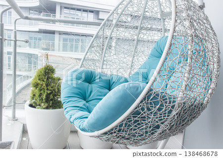 Cozy modern apartment concept. Cozy wicker hammock on balcony. Hanging chair with a blue pillow on wooden terrace. Luxury house design. Dream house decoration. Comfortable home with modern furniture. 138468678