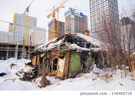 A dilapidated wooden house stands in a snowy landscape, overshadowed by towering cranes and modern buildings under construction in a bustling city. Reconstruction of the urban environment A dilapidated wooden house stands in a snowy landscape, overshadowed by towering cranes and modern buildings under construction in a bustling city. Reconstruction of the urban environment 138468729