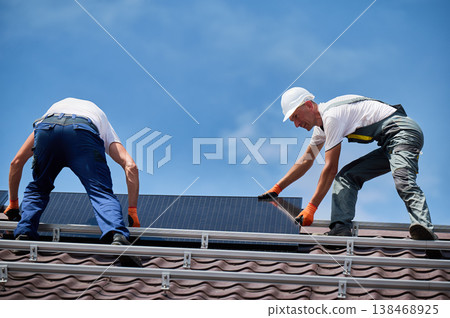 Workers building solar panel system on rooftop of house. Two men installers in helmets installing photovoltaic solar module outdoors. Alternative, green and renewable energy generation concept. Workers building solar panel system on rooftop of house. Two men installers in helmets installing photovoltaic solar module outdoors. Alternative, green and renewable energy generation concept. 138468925