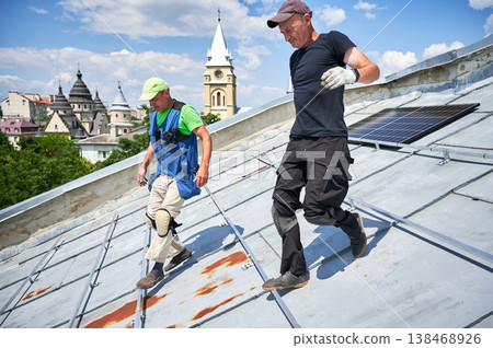 Workers building solar panel system on metal rooftop in urban setting. Two men installers carrying photovoltaic solar module outdoors. Alternative, green and renewable energy generation concept. 138468926