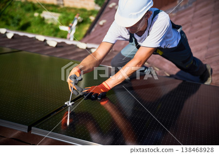 Worker building photovoltaic solar panel system on rooftop of house. Man engineer in helmets and gloves installing solar module with help of hex key outdoors. Alternative energy generation concept. Worker building photovoltaic solar panel system on rooftop of house. Man engineer in helmets and gloves installing solar module with help of hex key outdoors. Alternative energy generation concept. 138468928