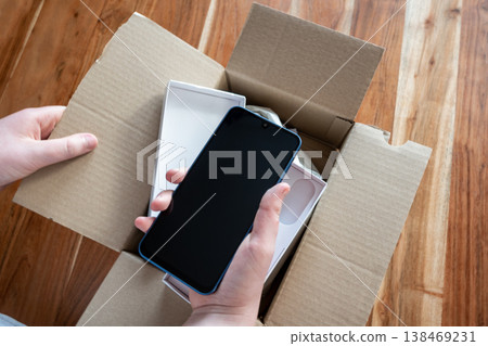 Man unpacking the cardboard box with new mobile phone on the wood table. Smartphone in the handsof a person. Man unpacking the cardboard box with new mobile phone on the wood table. Smartphone in the handsof a person. 138469231