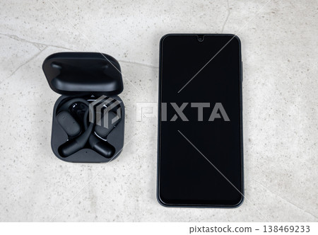 Black wireless in-ear headphones with case and mobile phone on the stone background. Flat lay. Earphones and smartphone. Black wireless in-ear headphones with case and mobile phone on the stone background. Flat lay. Earphones and smartphone. 138469233