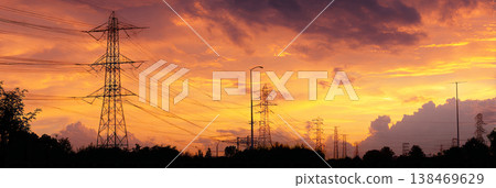High voltage power lines stretch across a wide field as the sun sets. The lines create a dark silhouette against the colorful sky filled with orange and blue hues High voltage power lines stretch across a wide field as the sun sets. The lines create a dark silhouette against the colorful sky filled with orange and blue hues 138469629