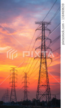 High voltage power lines deliver electricity to the city as night falls. The skyline is bright with lights from buildings and vehicles. Clouds hang over the scene creating contrast 138469639
