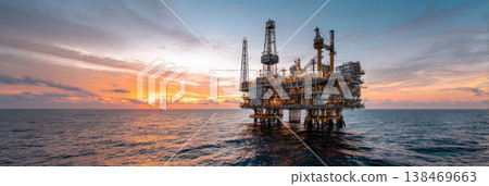 An oil platform stands tall above calm sea waters, with equipment and cranes visible. Bright sun lights up the scene, while clouds drift in the sky. The structure operates in the water 138469663