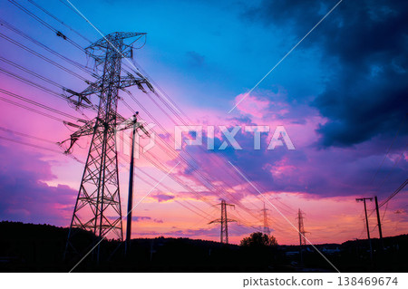 High voltage power lines stretch across a wide field as the sun sets. The lines create a dark silhouette against the colorful sky filled with orange and blue hues High voltage power lines stretch across a wide field as the sun sets. The lines create a dark silhouette against the colorful sky filled with orange and blue hues 138469674