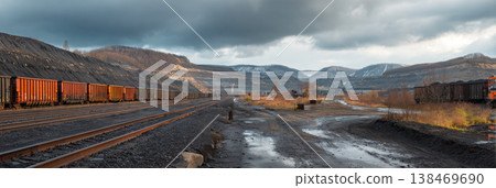 Coal mine shows a large spoil tip with rail cars positioned along the tracks. The scene features machinery and mining activity in the background with a cloudy sky Coal mine shows a large spoil tip with rail cars positioned along the tracks. The scene features machinery and mining activity in the background with a cloudy sky 138469690
