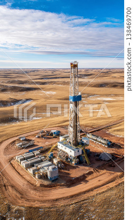 A drilling rig stands tall in a rural area with surrounding outbuildings and tanks. The sky is clear, showing a typical workday in oil or natural gas extraction 138469700