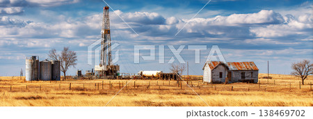 A drilling rig stands tall in a rural area with surrounding outbuildings and tanks. The sky is clear, showing a typical workday in oil or natural gas extraction 138469702