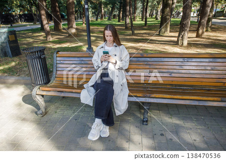 Nice woman using smartphone and sitting on bench outdoors Nice woman using smartphone and sitting on bench outdoors 138470536