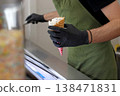 Worker holding ice cream cone with topping at counter in cafe. Food service scene for dessert preparation, retail work, sweet snack serving and professional kitchen routine. 138471831