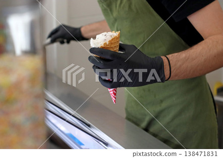 Worker holding ice cream cone with topping at counter in cafe. Food service scene for dessert preparation, retail work, sweet snack serving and professional kitchen routine. Worker holding ice cream cone with topping at counter in cafe. Food service scene for dessert preparation, retail work, sweet snack serving and professional kitchen routine. 138471831