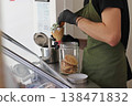 Worker preparing ice cream cone with scoop near container and waffle biscuits at counter. Dessert making process in cafe, food service work, sweet product preparation and retail environment. 138471832