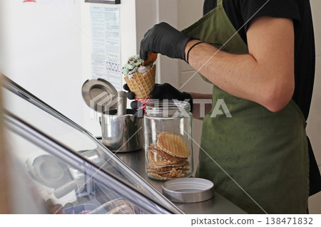 Worker preparing ice cream cone with scoop near container and waffle biscuits at counter. Dessert making process in cafe, food service work, sweet product preparation and retail environment. 138471832